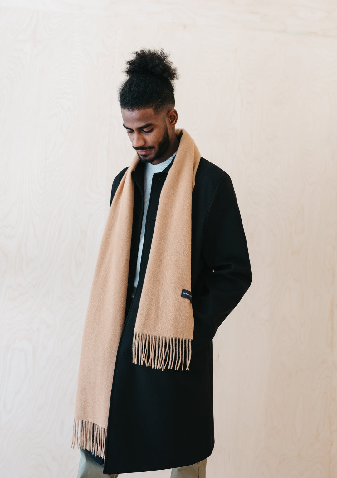 A person wearing TBCo Men's Lambswool Scarf in Camel draped elegantly over a black winter coat, showcasing the scarf's fringed detail