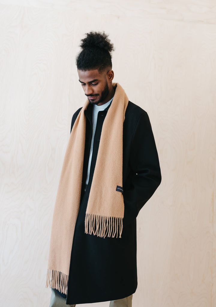A person wearing TBCo Men's Lambswool Scarf in Camel draped elegantly over a black winter coat, showcasing the scarf's fringed detail