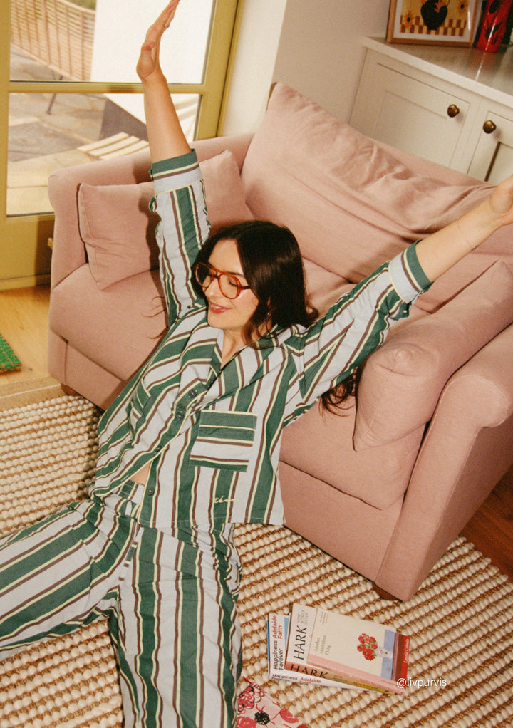 Woman stretching wearing TBCo Cotton Pyjamas in Blue Stripe sitting by a pink sofa in a cosy living room