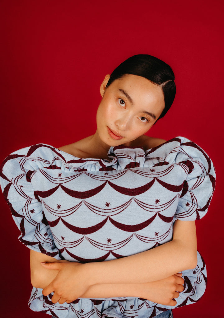 Young woman holding TBCo Cotton Cushion Cover in Blue Circus; features pale blue with maroon scallop and star pattern, oversized print, and ruffle edging.