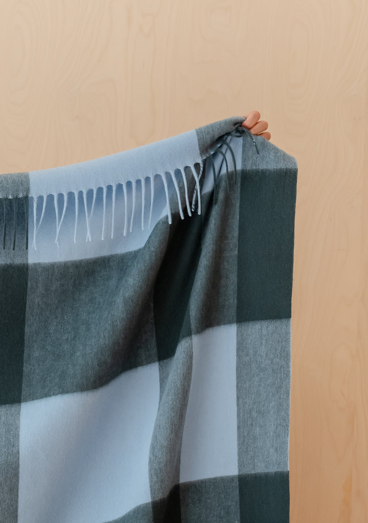 Hand holding TBCo Lambswool Blanket in Green Modern Check with fringed edges against wood background
