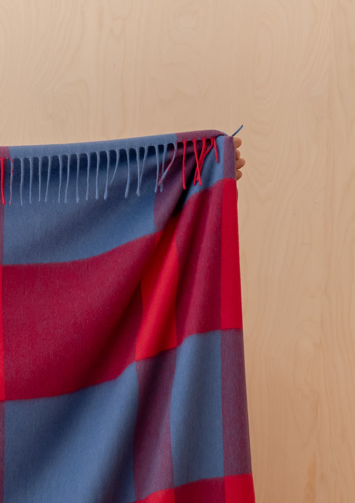 Handheld TBCo Lambswool Blanket in Red Modern Check with fringed edges against a light wooden background