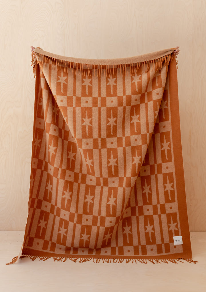 Person holding TBCo Lambswool Blanket in Almond Celestial Tile Jacquard with fringed edges against wooden background