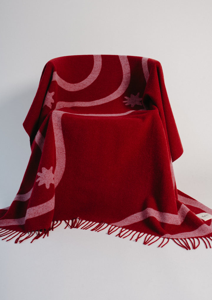 Lambswool Blanket in Red Wave Jacquard by TBCo draped over a chair showcasing its fringed edges and wave pattern
