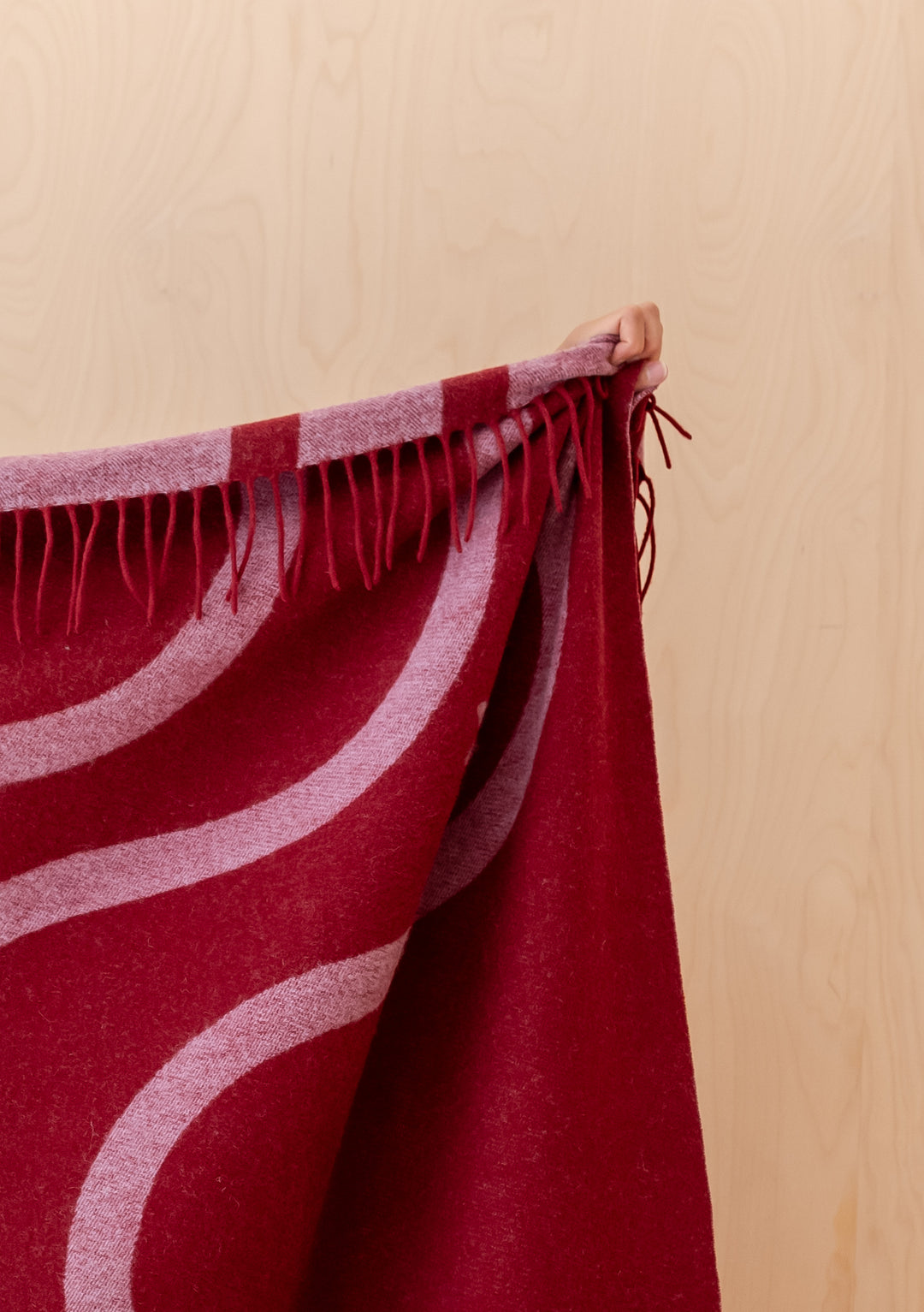 Hand holding fringed edge of TBCo Lambswool Blanket in Red Wave Jacquard with red and rose pink wave pattern
