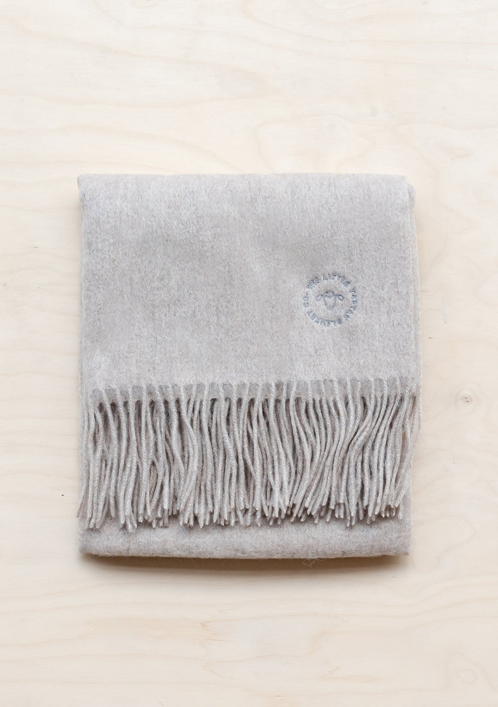 TBCo Lambswool Baby Blanket in Oatmeal Melange with fringed edge and embroidered logo, folded neatly on cream surface