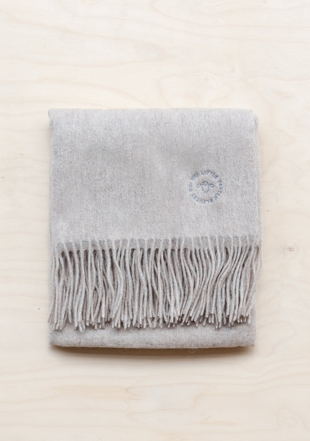 TBCo Lambswool Baby Blanket in Oatmeal Melange with fringed edge and embroidered logo, folded neatly on cream surface