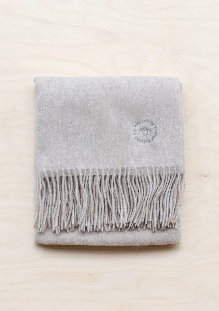 TBCo Lambswool Baby Blanket in Oatmeal Melange with fringed edge and embroidered logo, folded neatly on cream surface