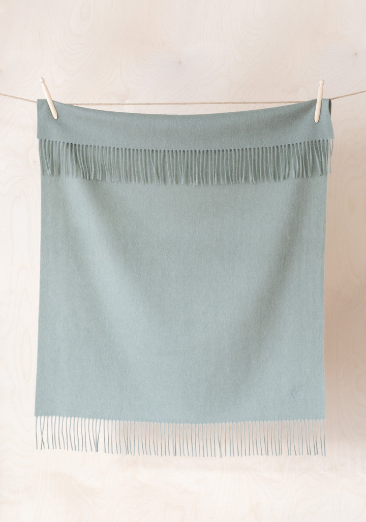 TBCo Lambswool Baby Blanket in Sage Melange hanging on clothesline, featuring soft fringe detail and gentle sage green colour