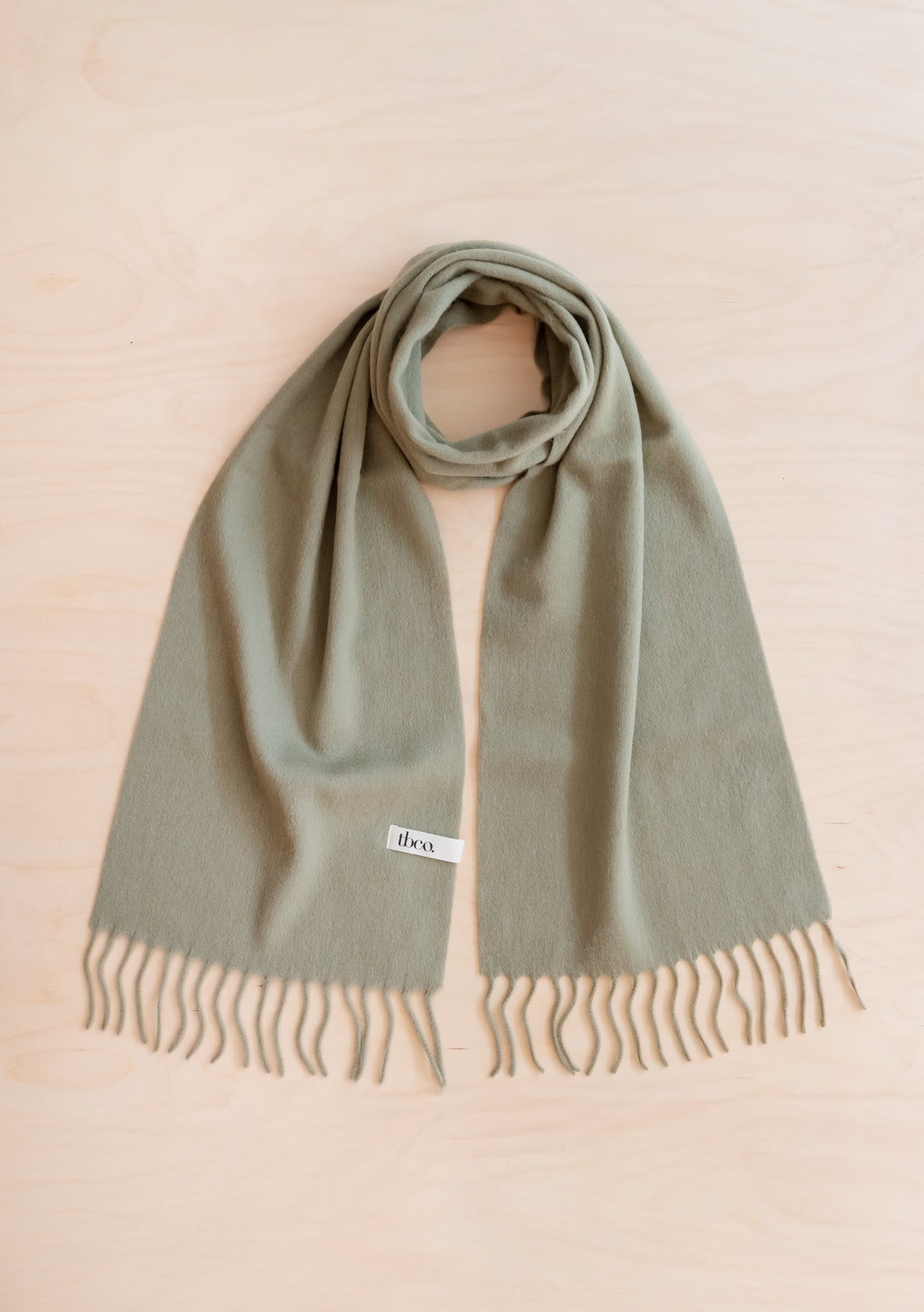 Lambswool Scarf in Olive by TBCo displayed flat with fringed edges on light wood background