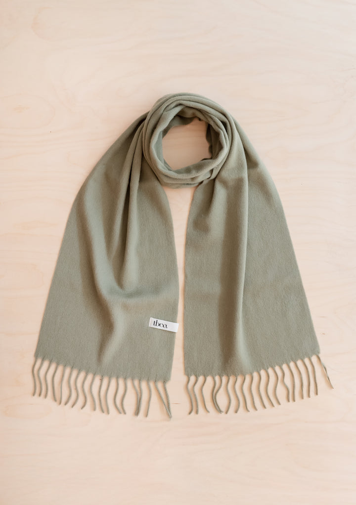 Lambswool Scarf in Olive by TBCo displayed flat with fringed edges on light wood background