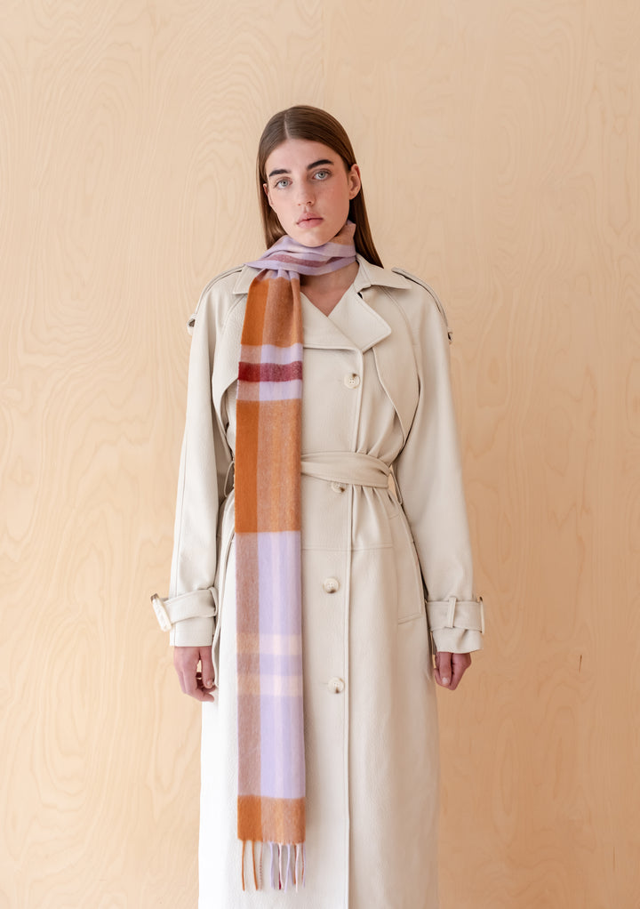 Model wearing a TBCo Lambswool Skinny Scarf in Lilac Modern Tartan with a beige trench coat against wood panel background