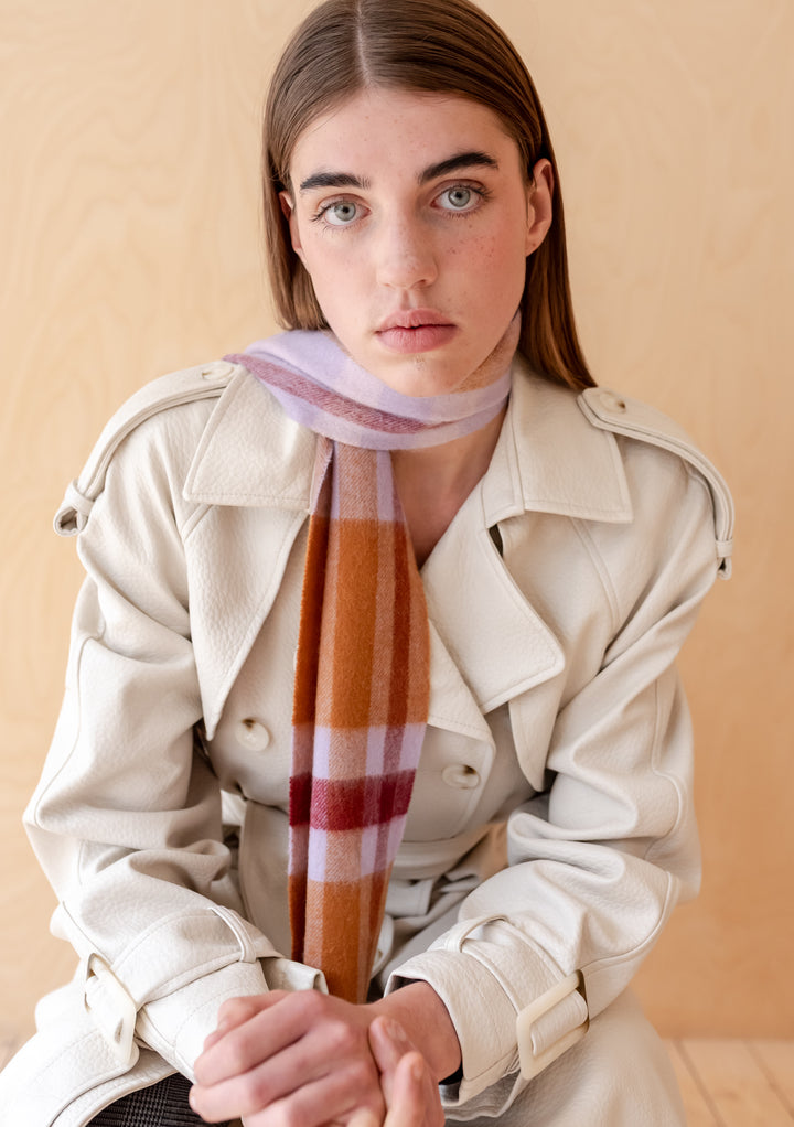 Woman wearing a TBCo Lambswool Skinny Scarf in Lilac Modern Tartan styled with a cream trench coat against a neutral background