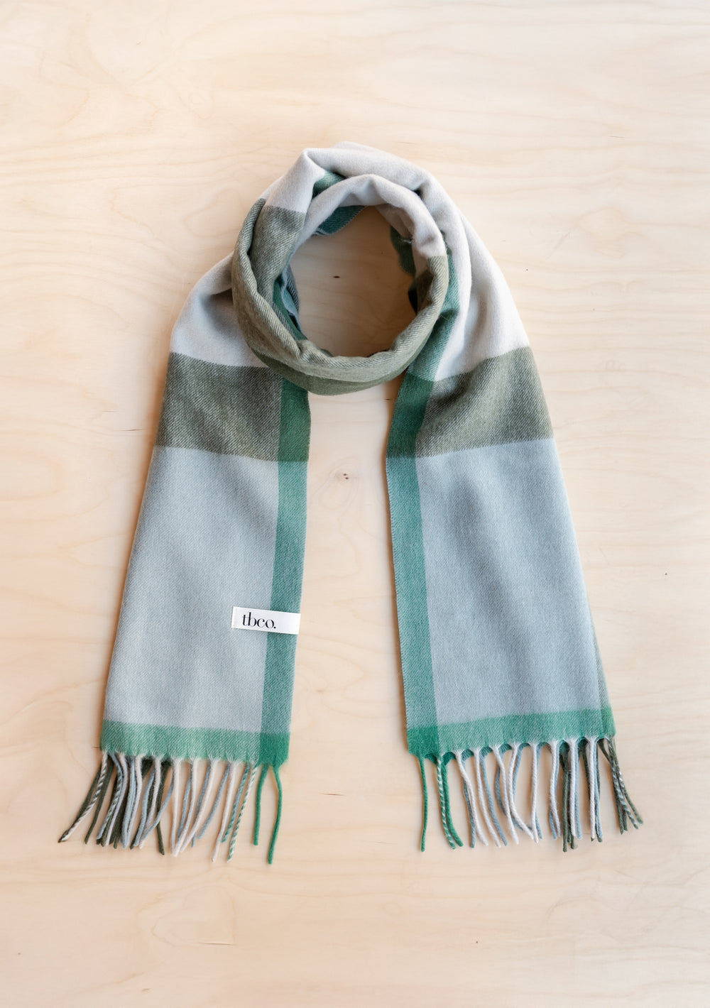 TBCo Lambswool Oversized Scarf in Green Block Check with fringed edges laid flat on wooden surface