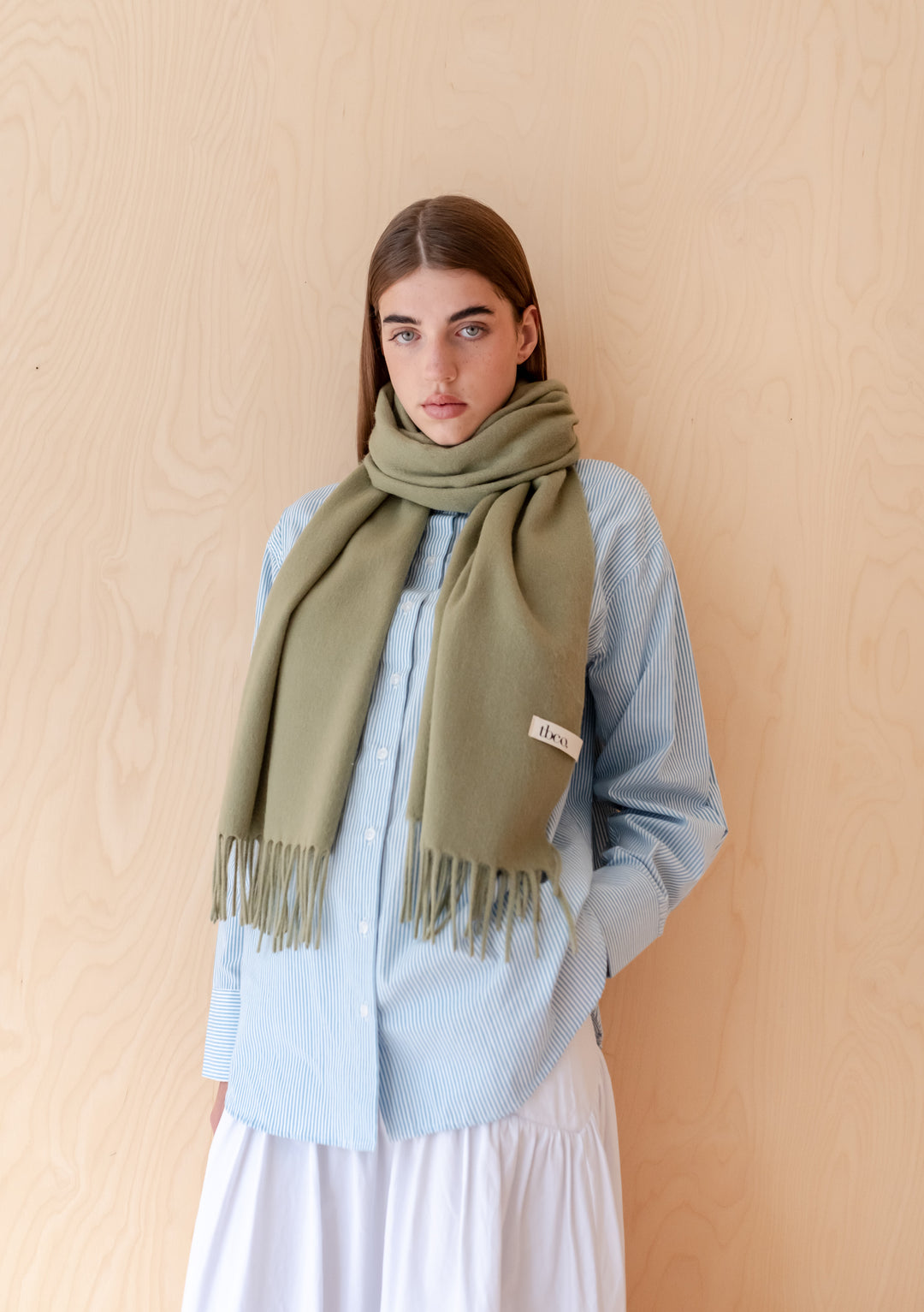 Model wearing a soft Lambswool Oversized Scarf in Olive by TBCo with fringed edges against wooden background
