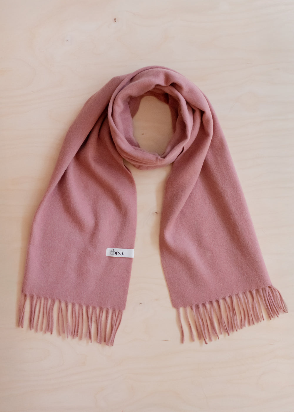 Soft Lambswool Oversized Scarf in Blush by TBCo with fringed edges, laid flat on light wood background