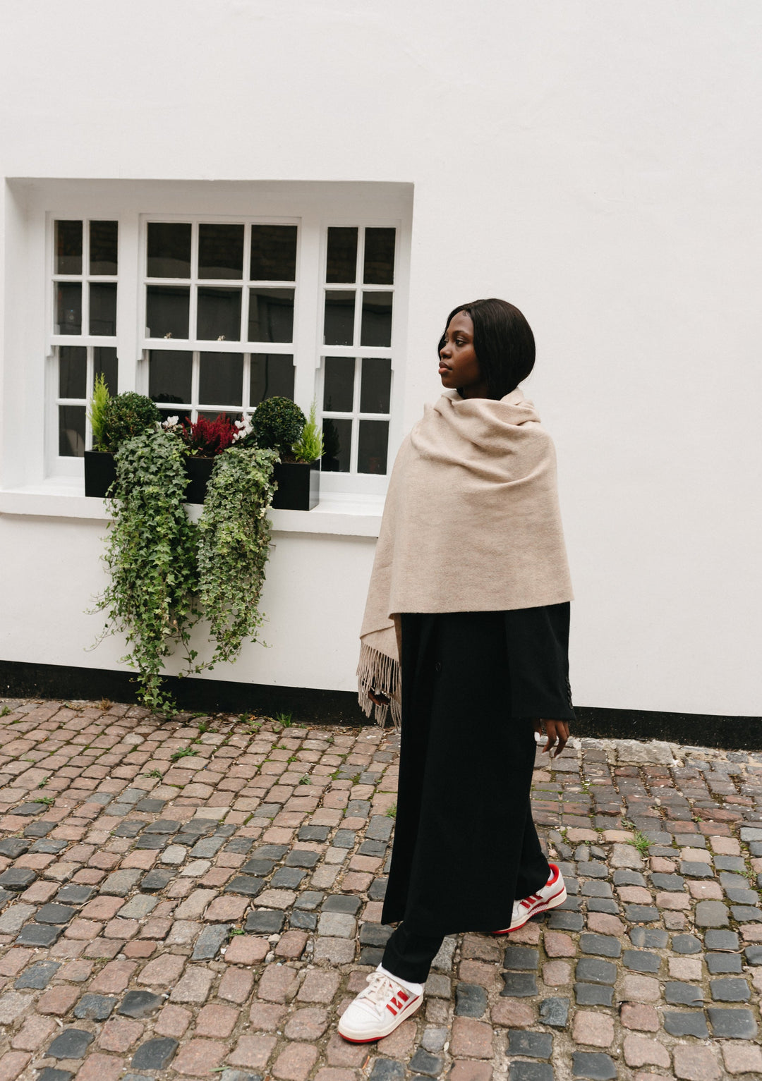 Elegant TBCo Lambswool Blanket Scarf in Oatmeal Melange styled with a black dress on cobblestone street beside white wall with window box