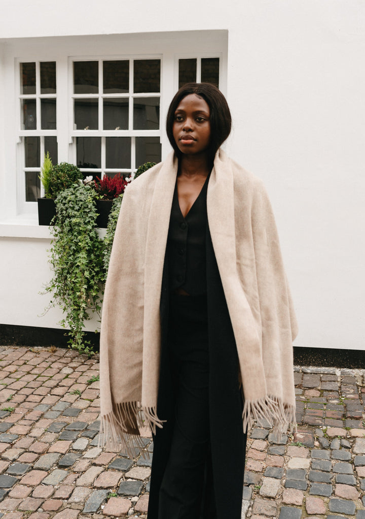 Luxurious TBCo lambswool blanket scarf in oatmeal melange styled with black outfit on cobblestone courtyard with white building
