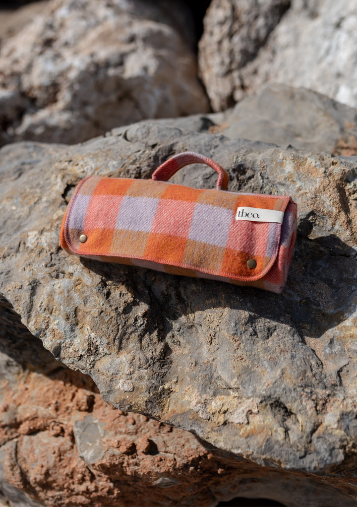 TBCo Recycled Wool Small Picnic Blanket in orange gingham pattern, rolled and secured with leather handle, displayed on rocky surface
