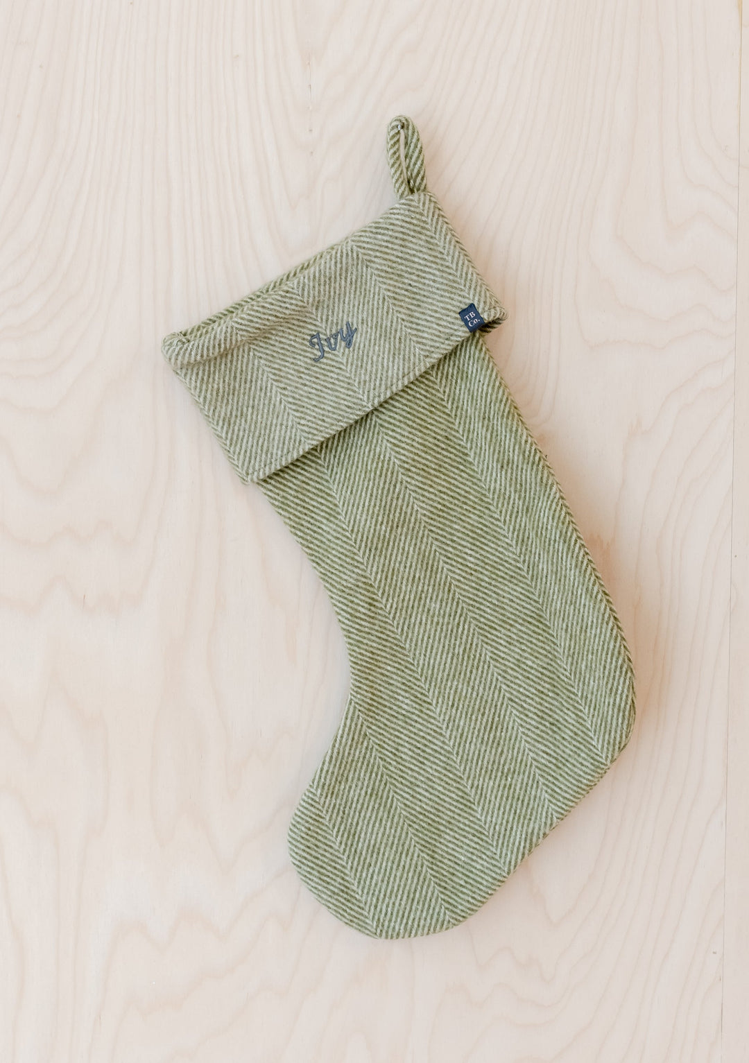 Sage green herringbone wool Christmas stocking with custom TBCo embroidery service available, displayed on light wood background