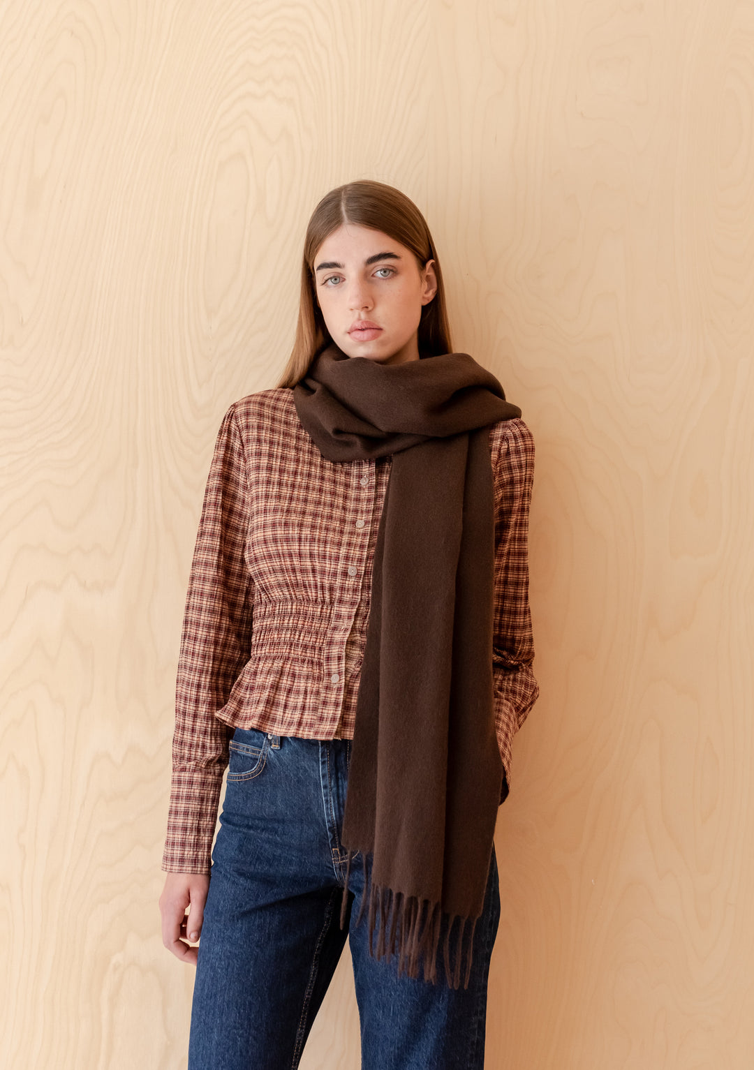 Model wearing TBCo Lambswool Oversized Scarf in Chocolate with fringed edges and autumn outfit against wooden background