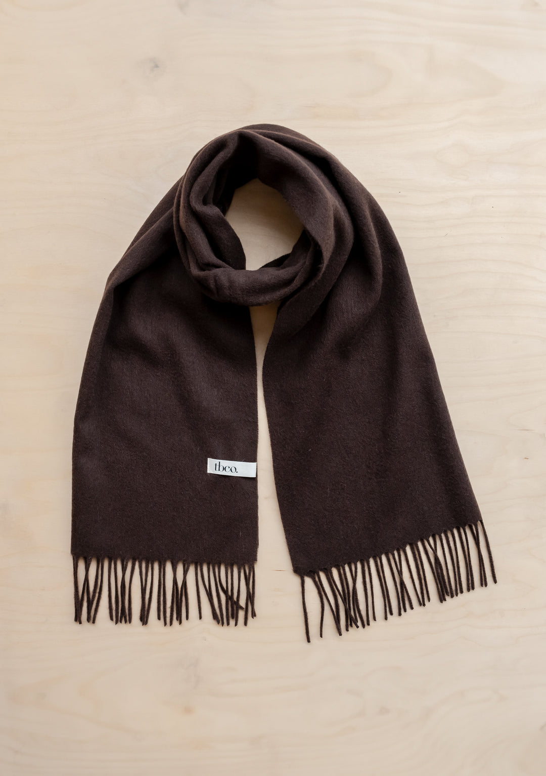 TBCo lambswool oversized scarf in chocolate brown with fringed edges, styled flat against wooden surface