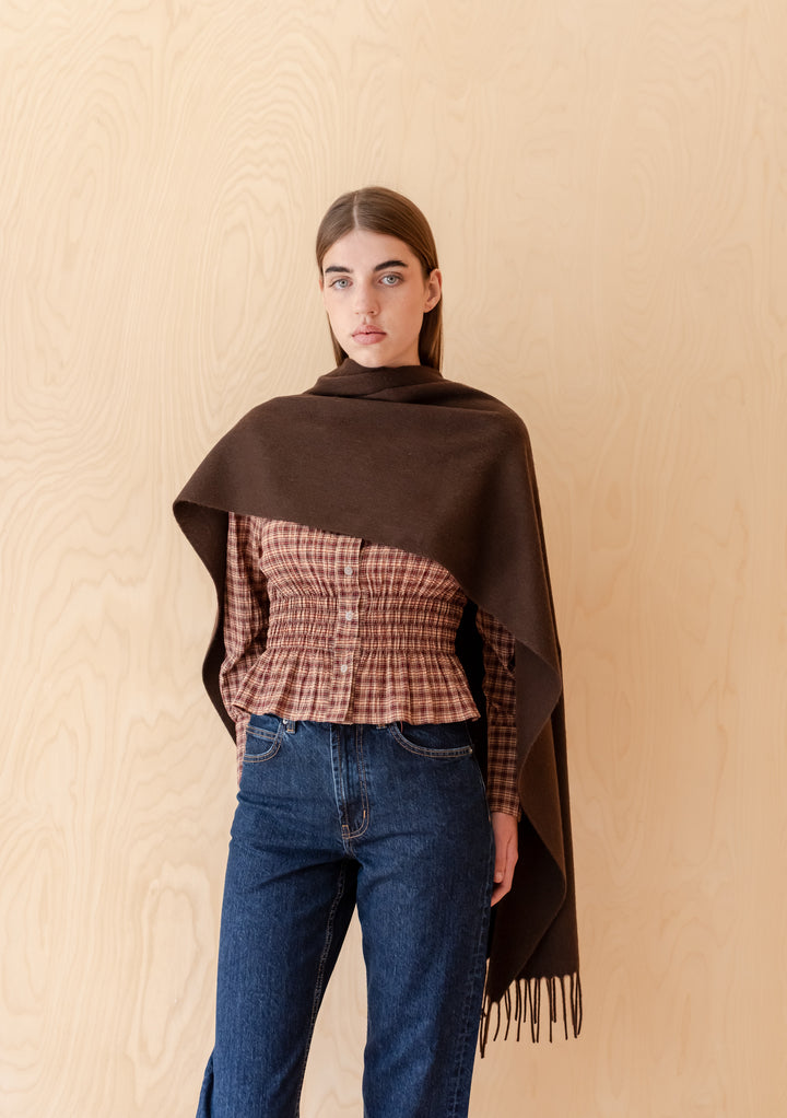 Model wearing TBCo Lambswool Oversized Scarf in Chocolate with fringed edges against a neutral wood background