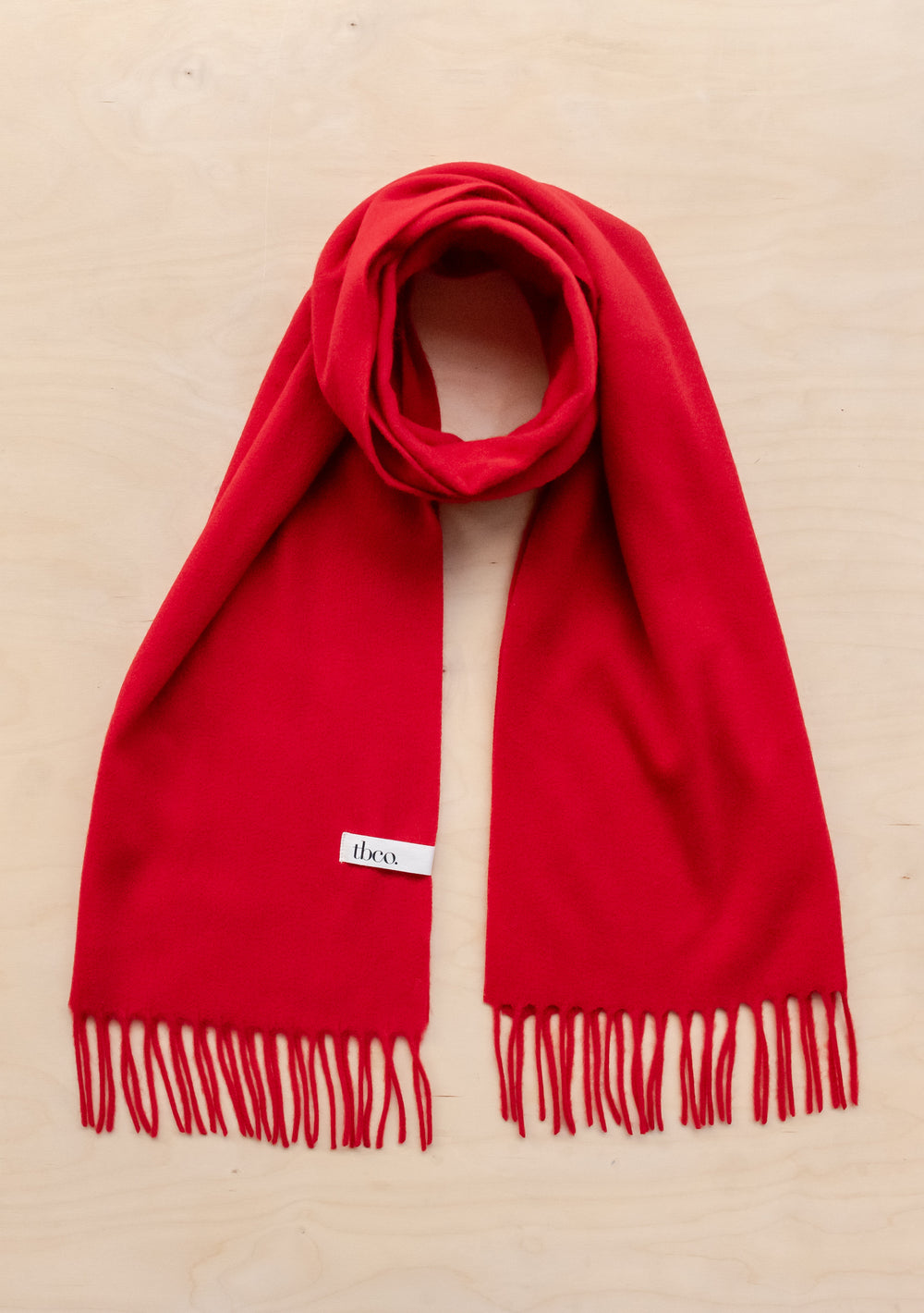 Bright red TBCo lambswool oversized scarf with fringed edges laid flat against beige background