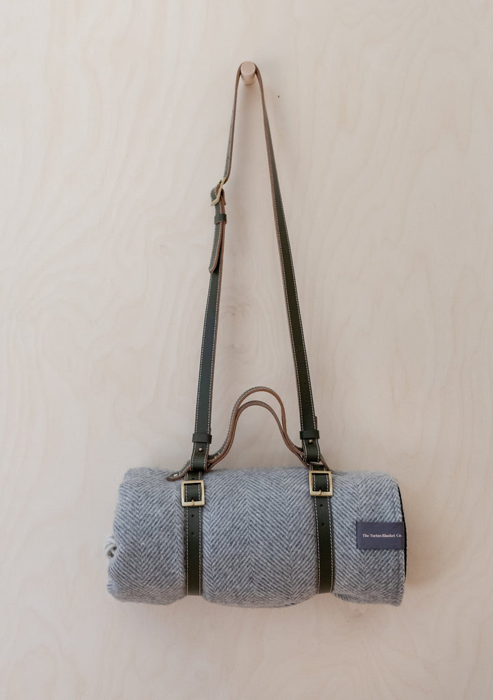 TBCo Leather Picnic Carrier with adjustable shoulder strap and brass buckles, carrying rolled grey herringbone wool blanket