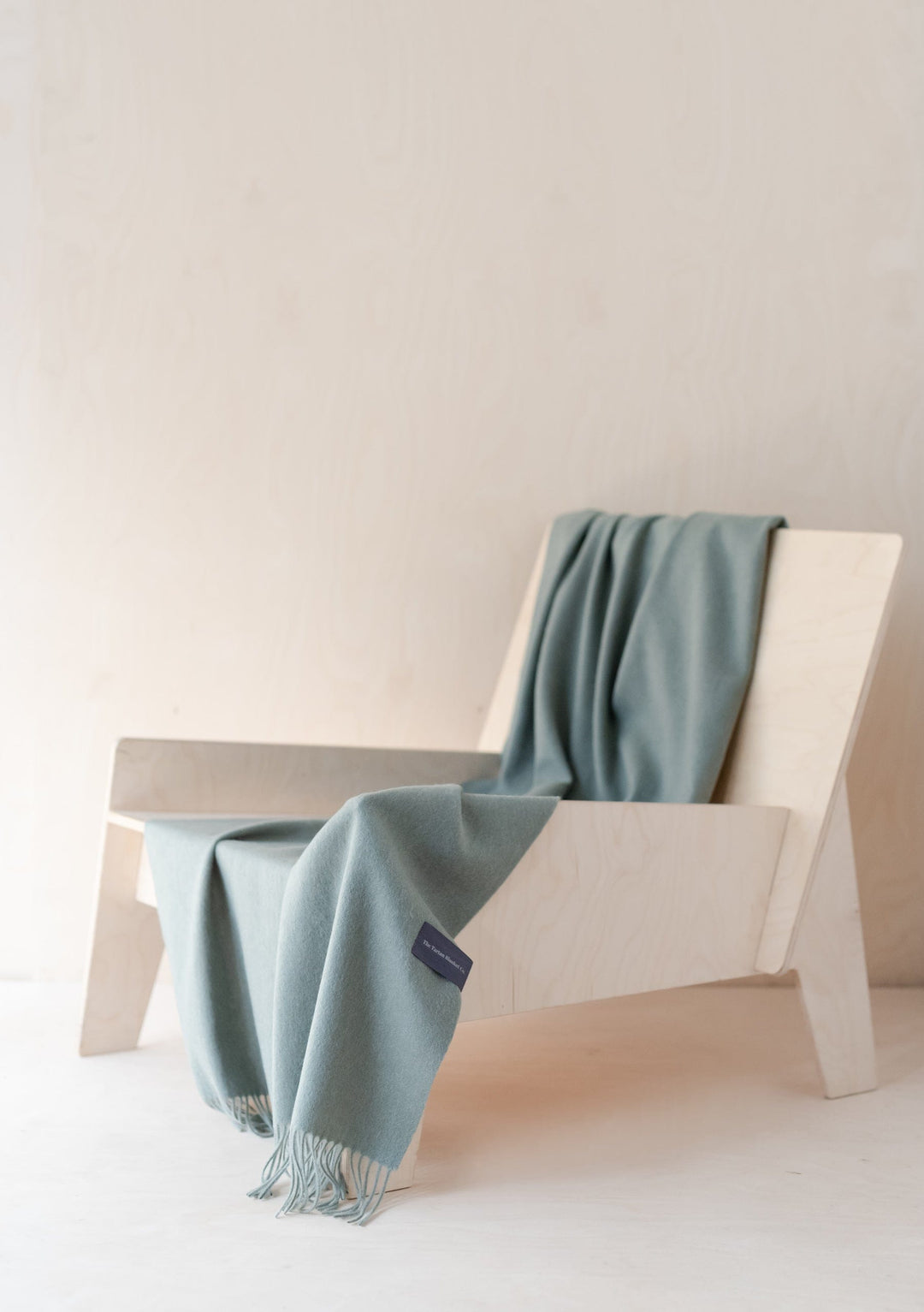 TBCo Lambswool Small Blanket in Sage Melange draped elegantly over white chair, showcasing soft texture and fringed detail