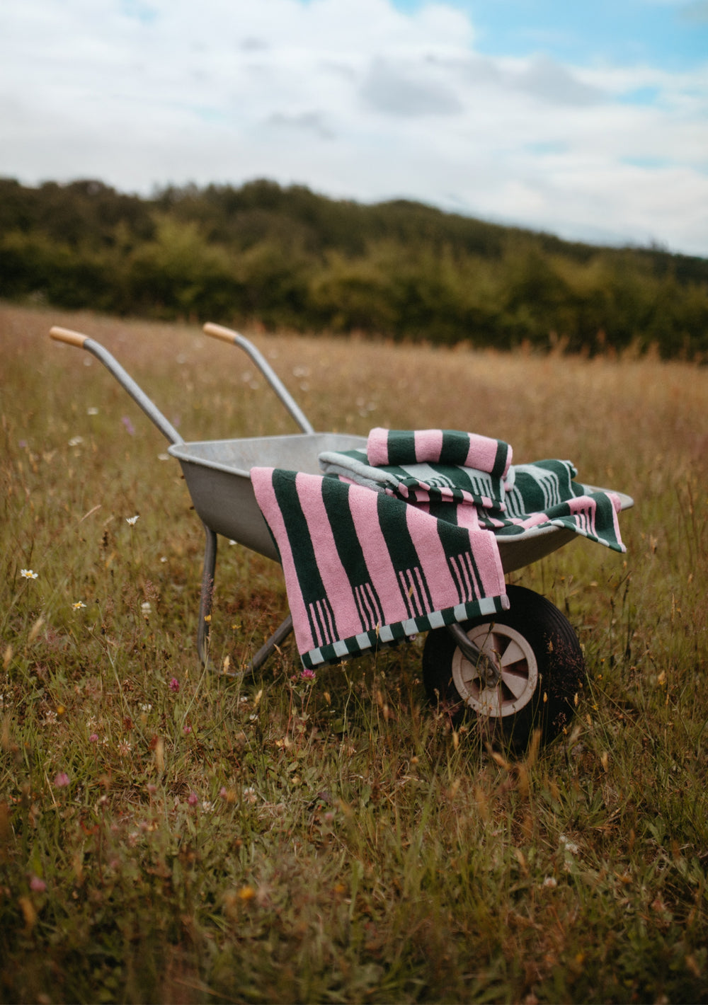 TBCo Terry Cotton Towels in Rose Stripe draped over a wheelbarrow in a grassy field with pink and green stripes.