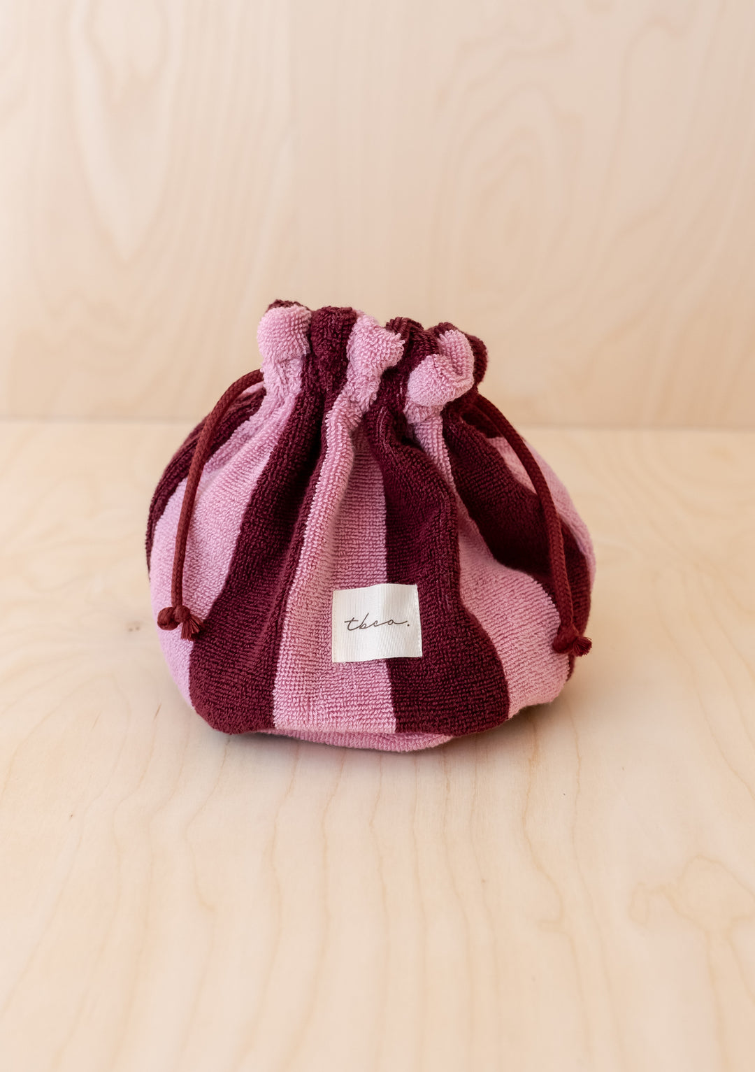The Terry Cotton Wash Bag in Burgundy Stripe, featuring pink and burgundy stripes and a drawstring top, rests on a light wooden surface. A white label with text is sewn onto the front of the small bag.