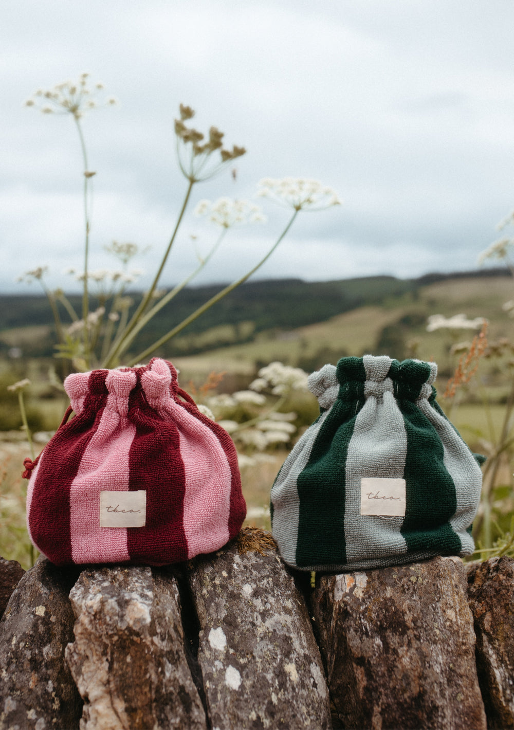 The Terry Cotton Wash Bag in Burgundy Stripe and a green-and-grey striped version rest on logs outdoors, with wildflowers and a blurred natural backdrop beneath a cloudy sky.