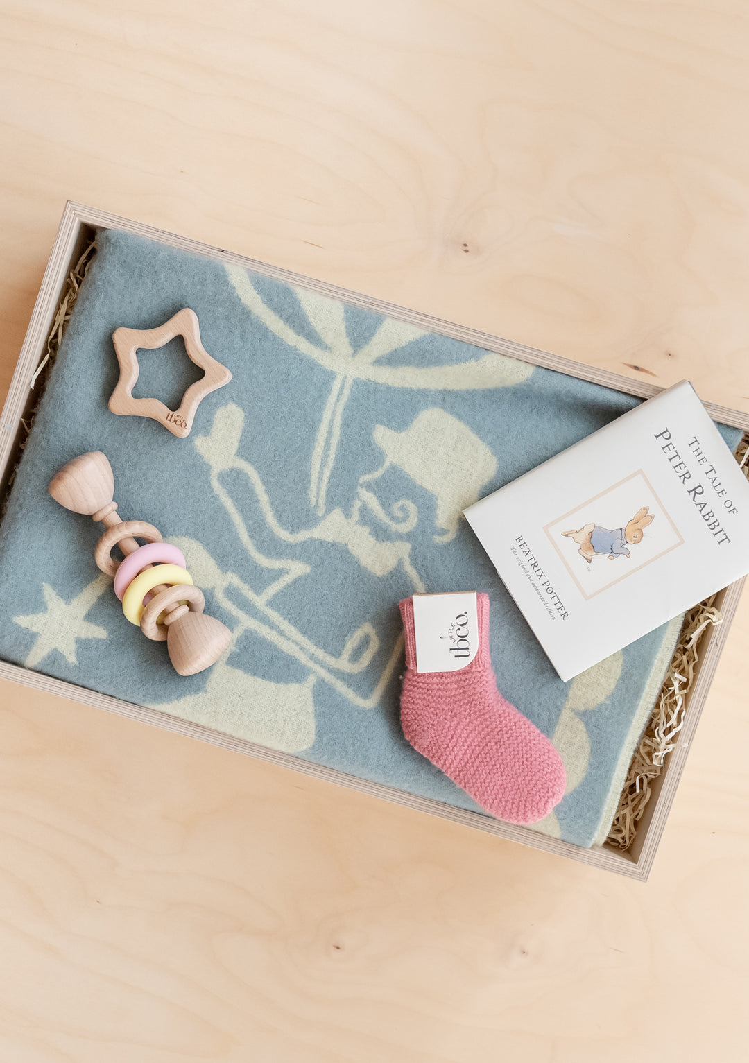 TBCo Baby Gift Box featuring a soft blanket, wooden toys, a pink sock, and a Peter Rabbit book for newborns.