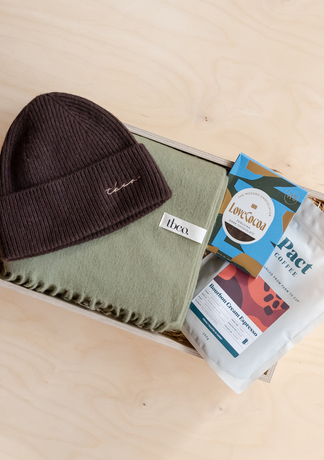 Gift Box For Him by TBCo featuring a brown beanie, green scarf, coffee pack, and chocolate bar in a wooden gift box.