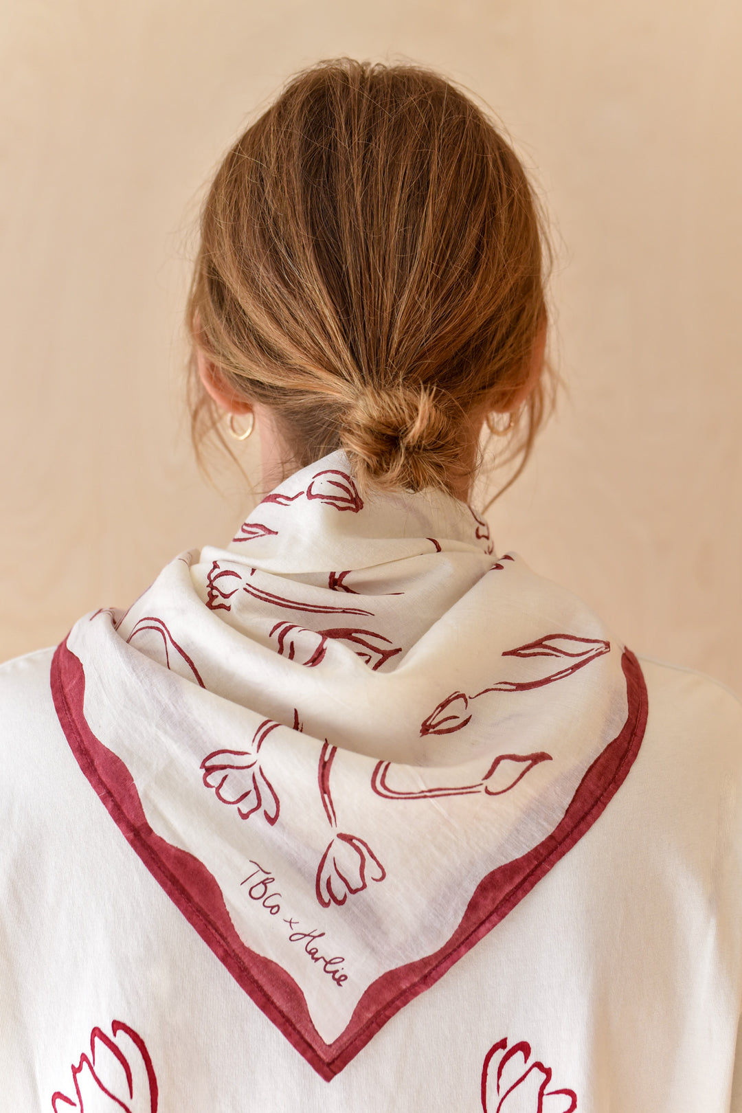 Close-up of a woman from behind wearing a TBCo x Harlie Briggs Artist Series cotton triangle neck scarf in white with red floral line art, featuring the TBCo x Harlie logo, styled over a low bun hairstyle against a soft beige background.
