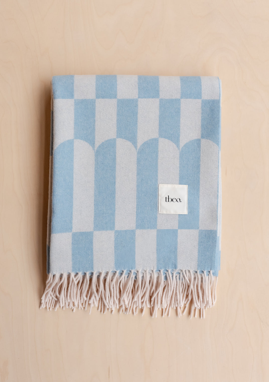 Folded Lambswool Blanket in Blue Geo by TBCo with fringe detail and soft blue geometric arch pattern on pale wood background