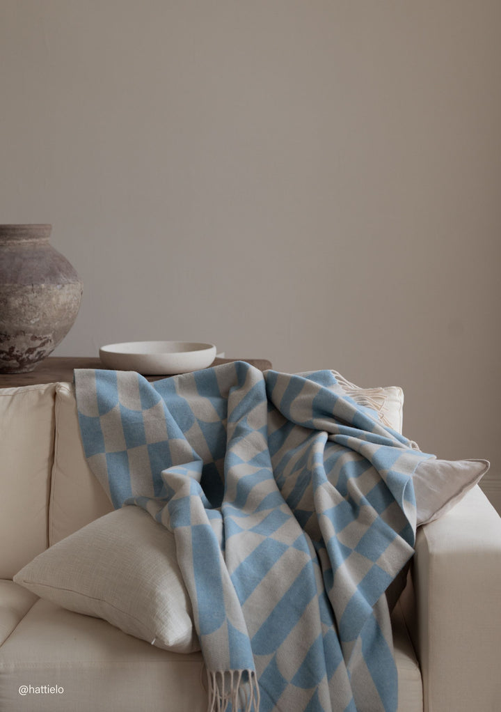 TBCo Lambswool Blanket in Blue Geo with soft fringes draped over cream sofa, showing checkered blue and beige pattern