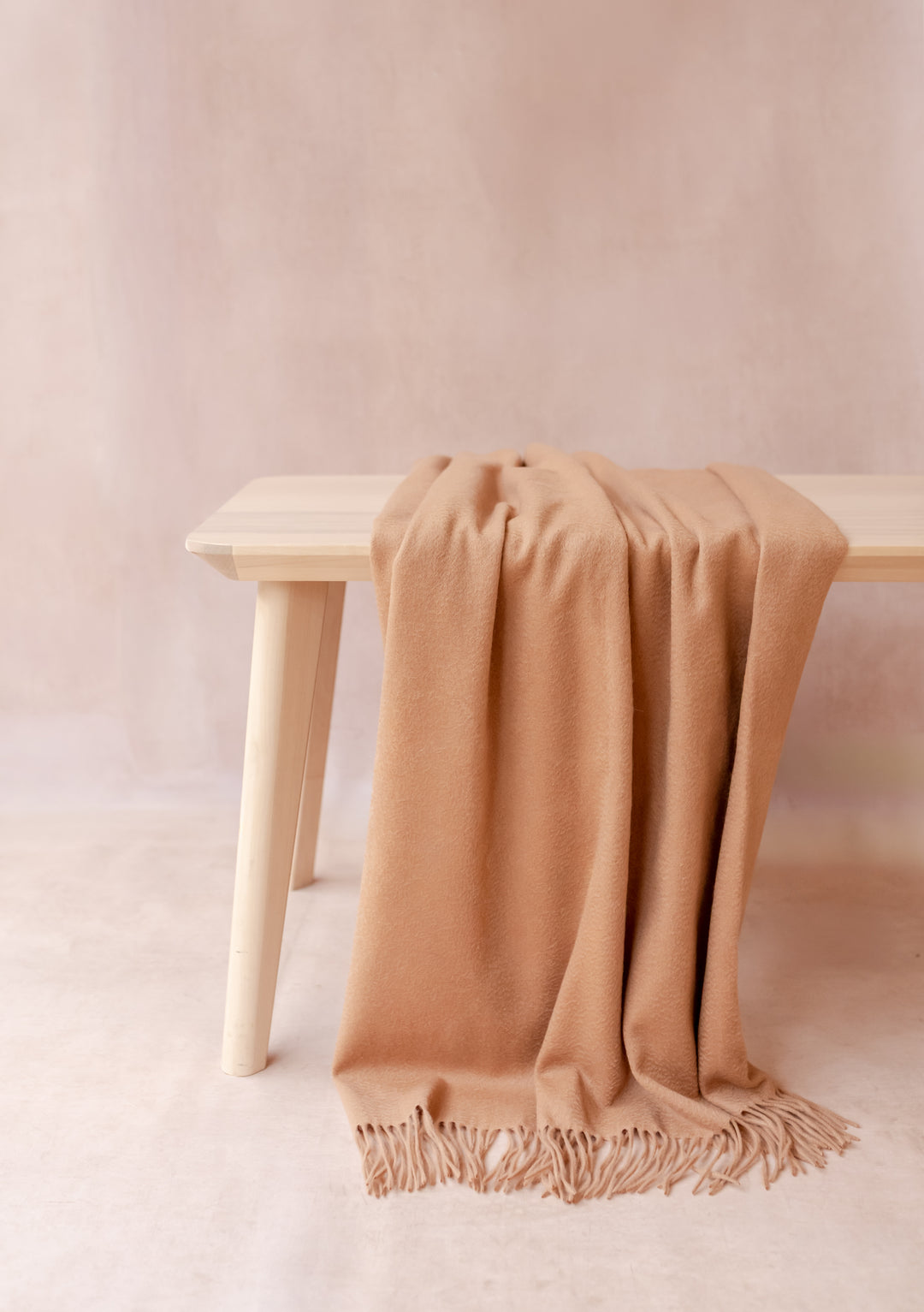 Soft camel cashmere small blanket with fringe by TBCo draped elegantly over a light wooden bench