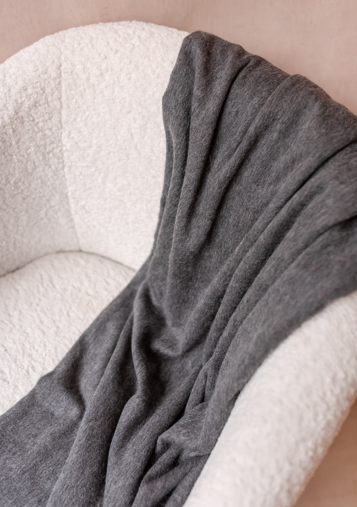 TBCo Cashmere Blanket in Charcoal Melange draped over a white textured armchair showcasing soft, luxurious fabric with fringe edges.