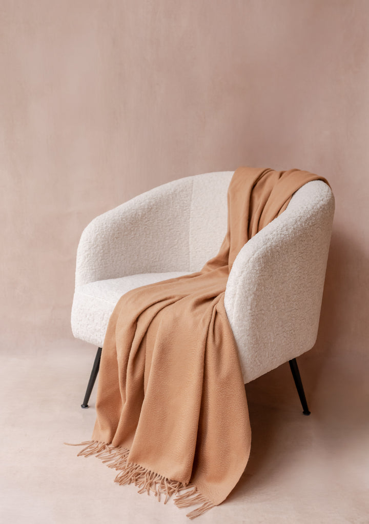 Cashmere Blanket in Camel by TBCo draped over a textured cream chair with soft fringe detailing