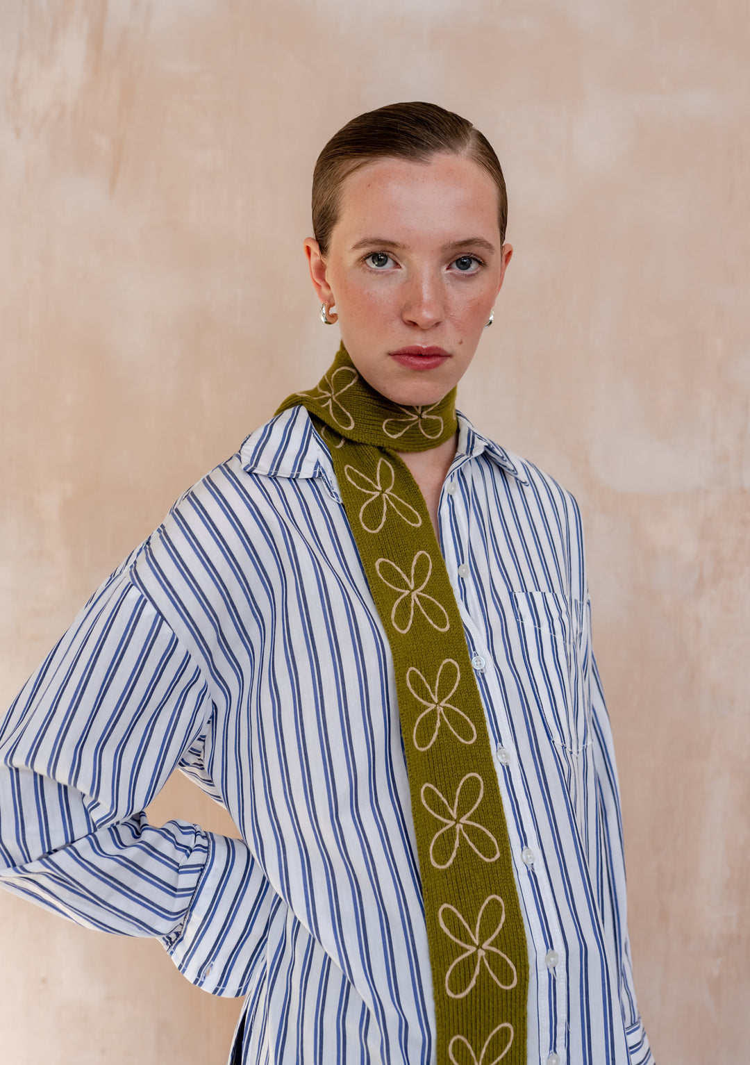 Close-up of Model wearing a TBCo Merino Wool Skinny Scarf in Green Flora, adorned with white floral embroidery, layered over a blue stripe shirt.
