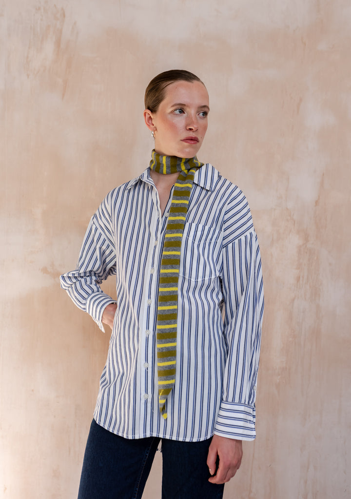 Model wearing a TBCo Merino Wool Neck Tie in Green Stripe, styled over a blue stripe shirt and dark jeans, showcasing its subtle elegance and transitional-inspired design.