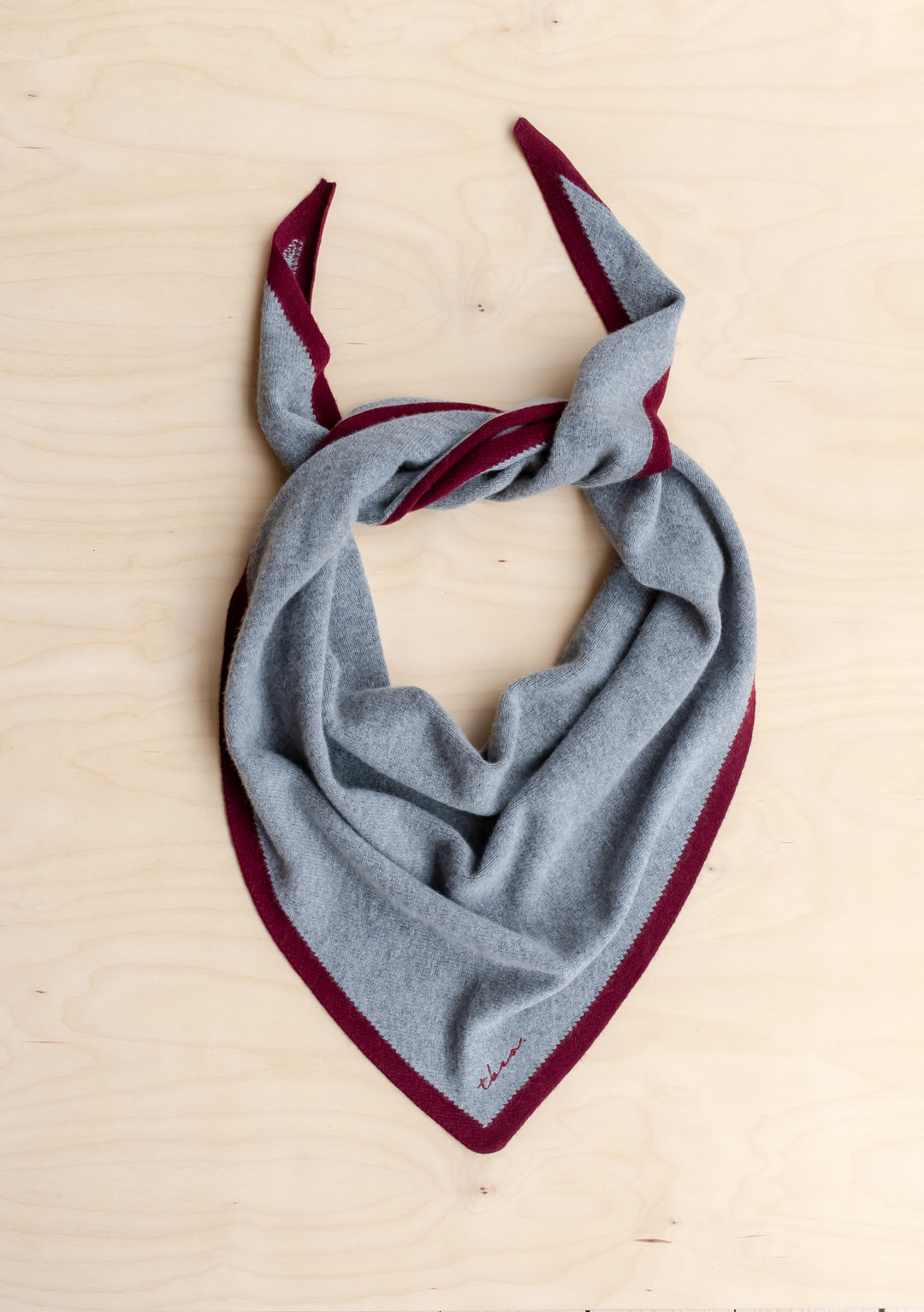 TBCo Large Merino Wool Triangle Scarf in Charcoal & Burgundy, featuring a light grey body, burgundy tipped border, and a loose knot on a wood surface.