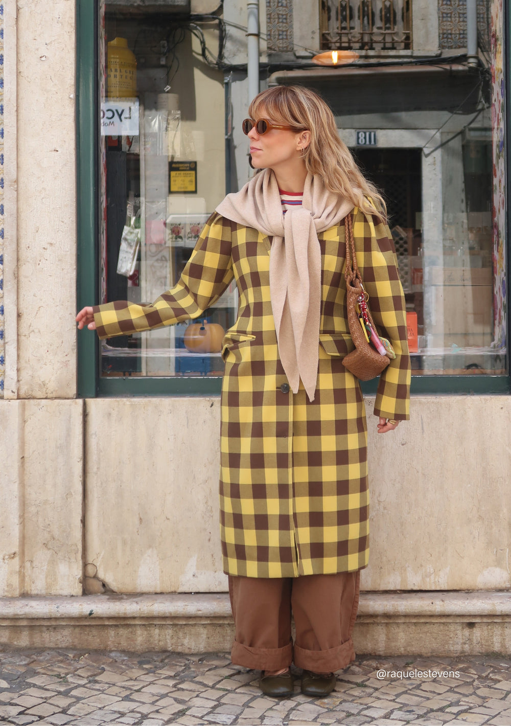Woman in checkered coat wearing TBCo Large Merino Wool Triangle Scarf in Oatmeal, styled draped on shoulders, in urban street setting.