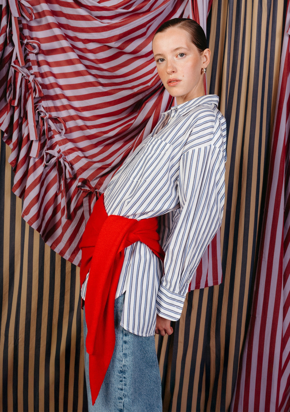 Model wearing a large merino triangle scarf in red by TBCo tied around waist over striped shirt and denim skirt