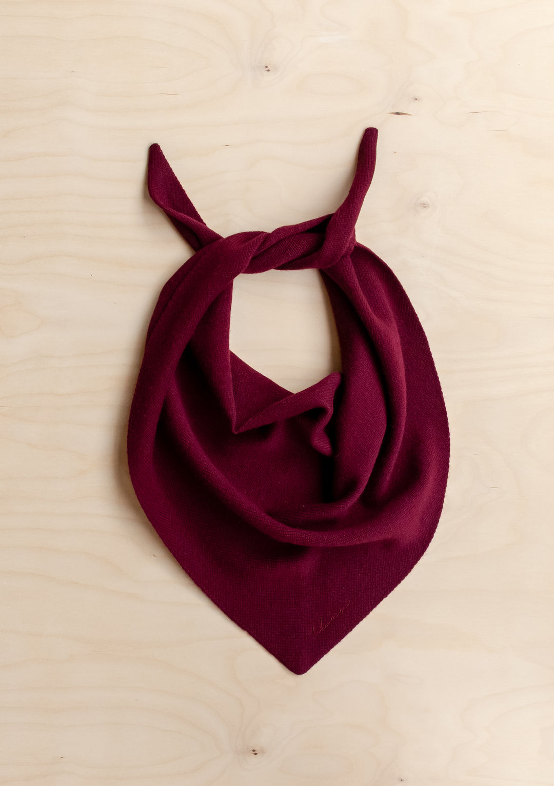 TBCo Merino Wool Triangle Scarf in Burgundy, flat on wooden surface, showcasing its embroidered TBCo logo and soft merino wool texture, ideal for versatile styling.