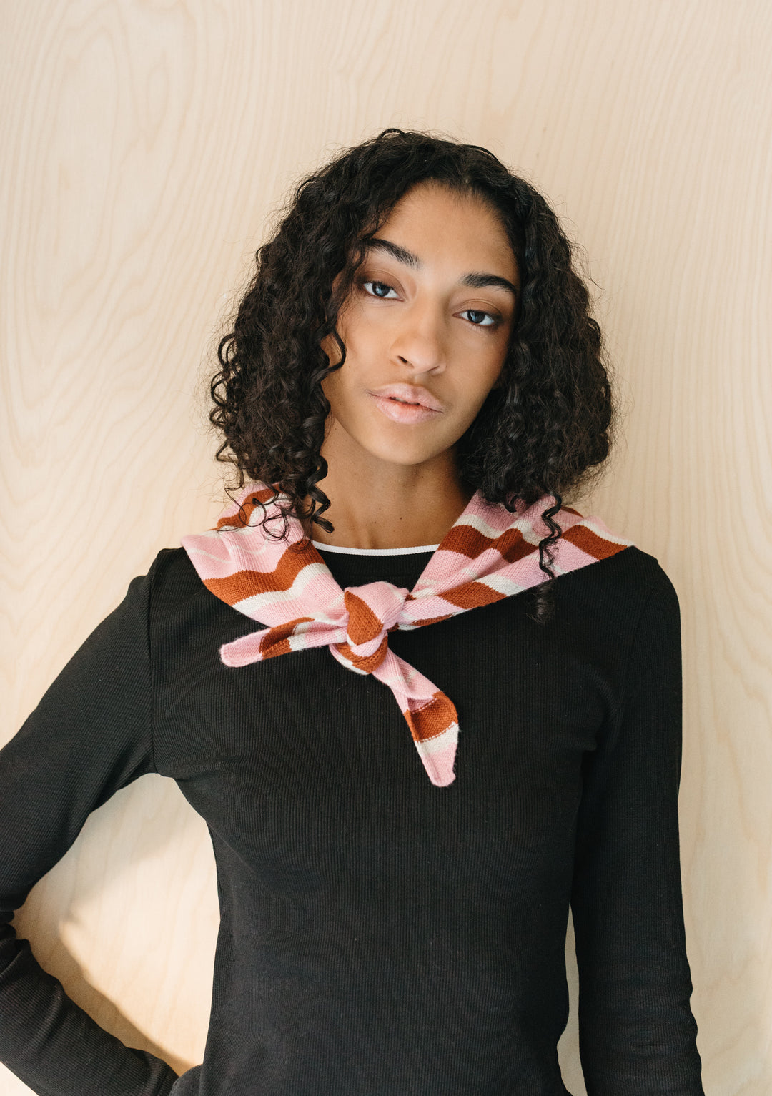 Model wearing TBCo Merino Wool Triangle Scarf in Pink & Rust Stripe tied at front over black top against wood background