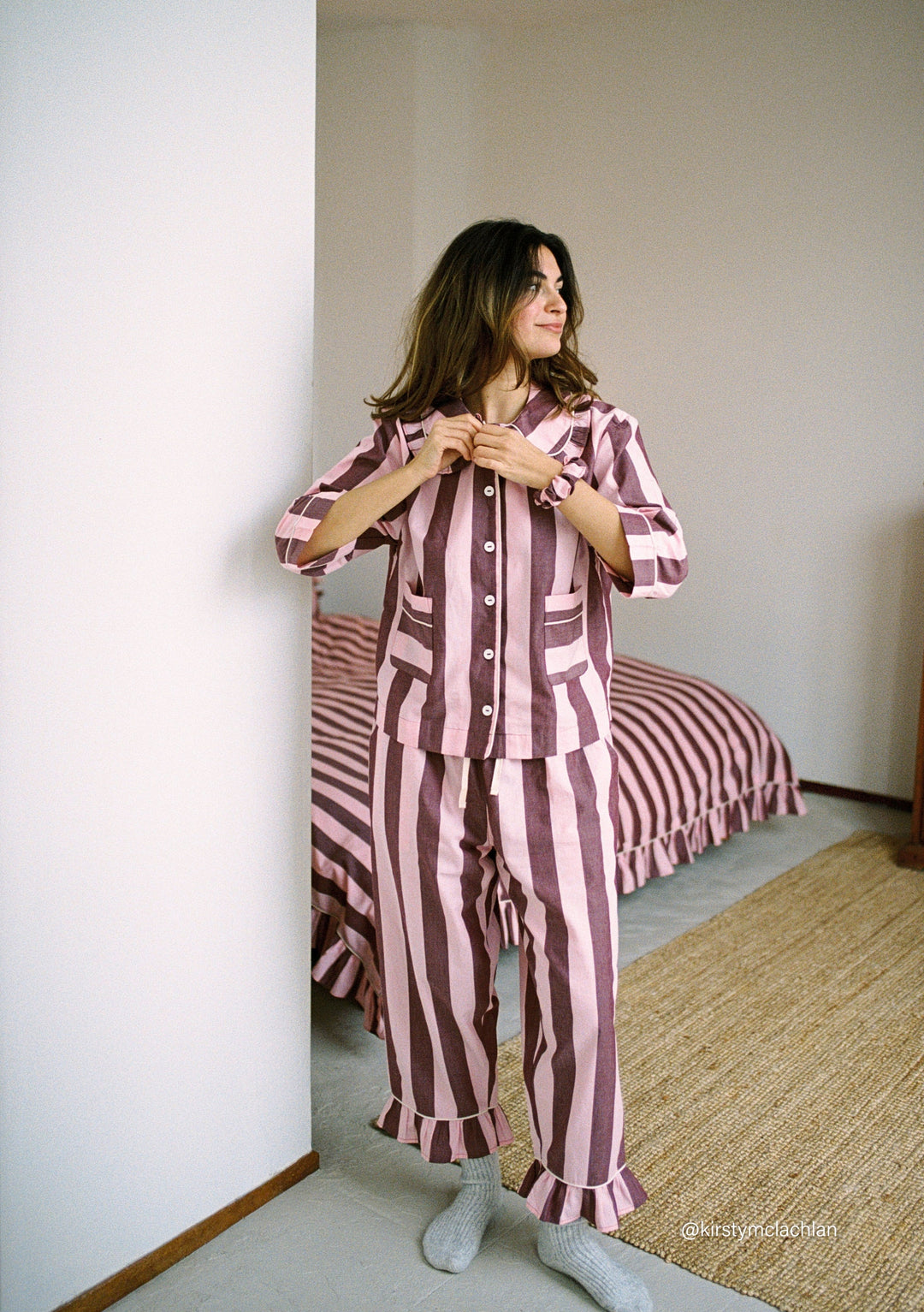 Woman wearing TBCo Cropped Cotton Pyjamas in Pink & Burgundy Stripe, standing by wall in cozy bedroom setting