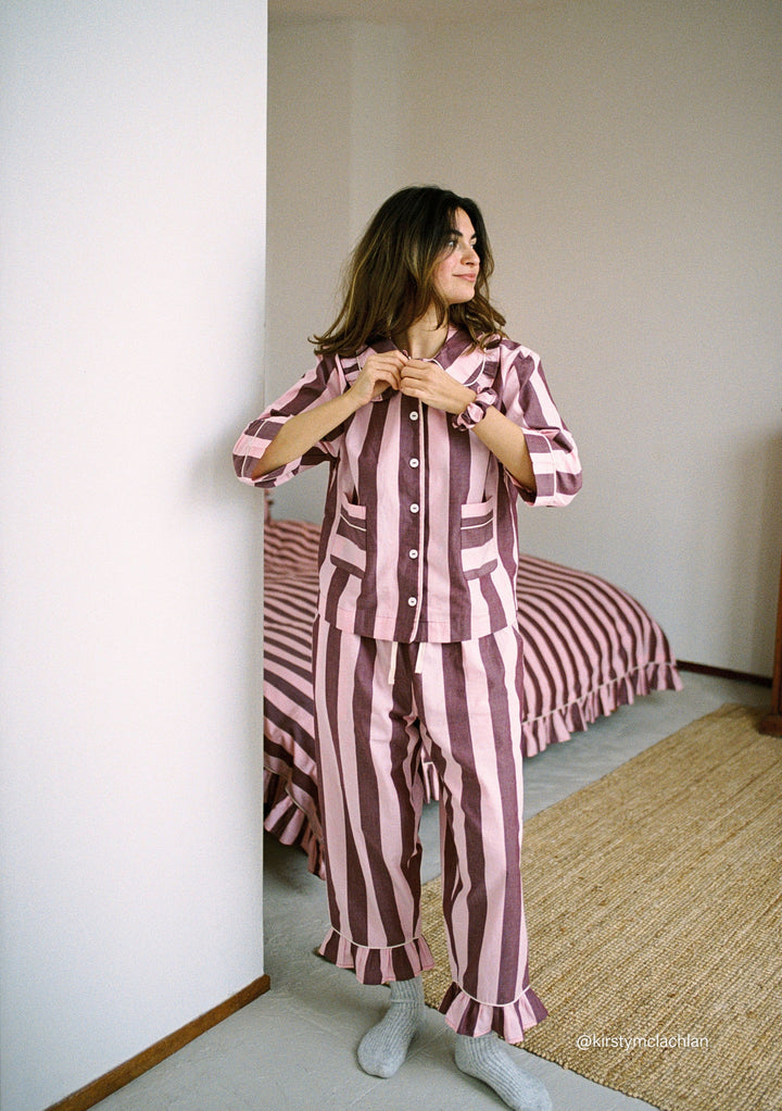 Woman wearing TBCo Cropped Cotton Pyjamas in Pink & Burgundy Stripe, standing by wall in cozy bedroom setting